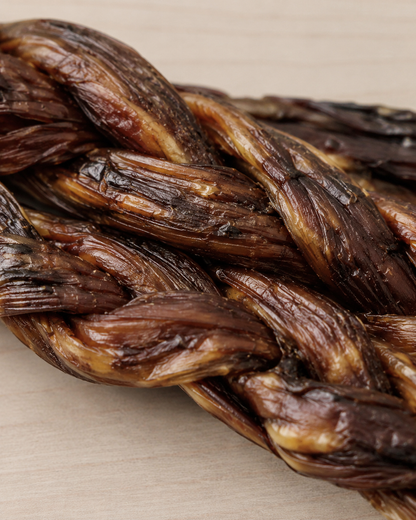 A close-up of a braided dog chew treat, showing the glossy, textured surface of the woven food on a light wood background.