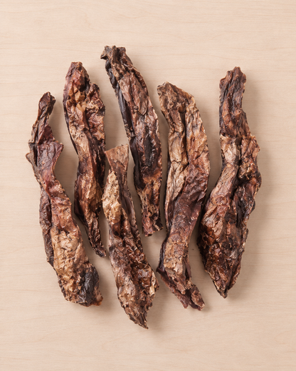 Five strips of dark brown jerky  arranged vertically on a light beige wood-grain surface.