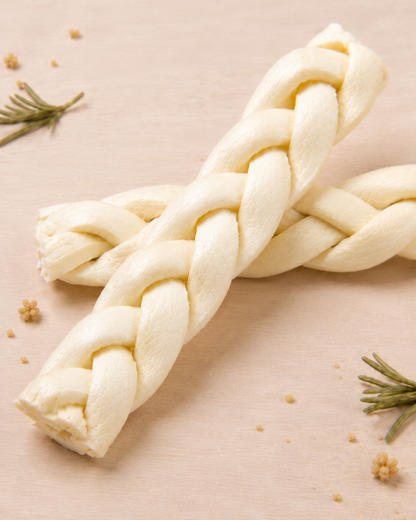 Allgood braided beef cheek, two  dog chews crossed in an X shape on a light beige surface with small rosemary sprigs and crumbs.