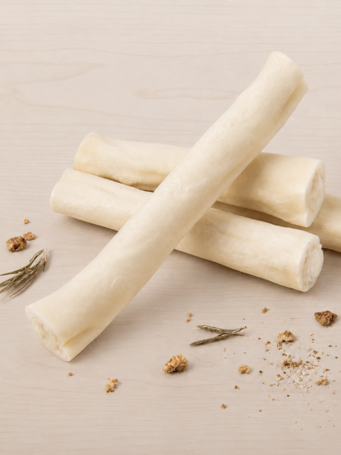 Four off-white bone-shaped dog treats arranged on a light wood surface with small scattered crumbs and herbs.