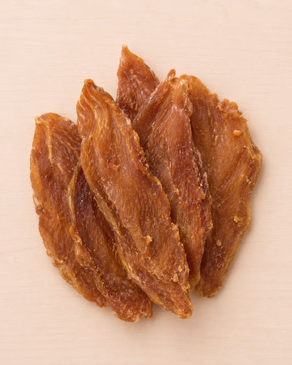 Four strips of chewy, golden-brown dried jerky arranged in a pile on a light beige wooden surface.