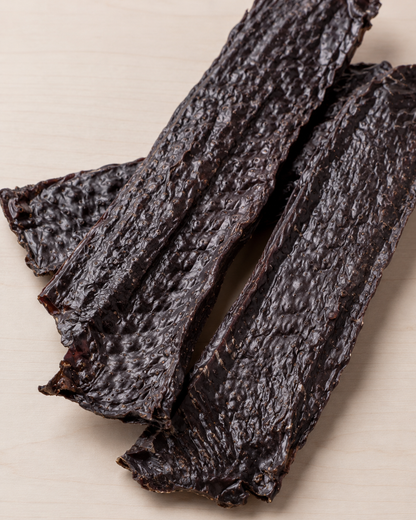 Three strips of dark, wrinkled jerky or dried meat snacks arranged diagonally on a light beige wooden surface.