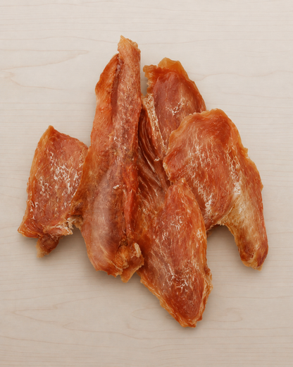 Four strips of dried, reddish-brown meat, likely chicken or turkey jerky, arranged on a light beige wood-grain surface.