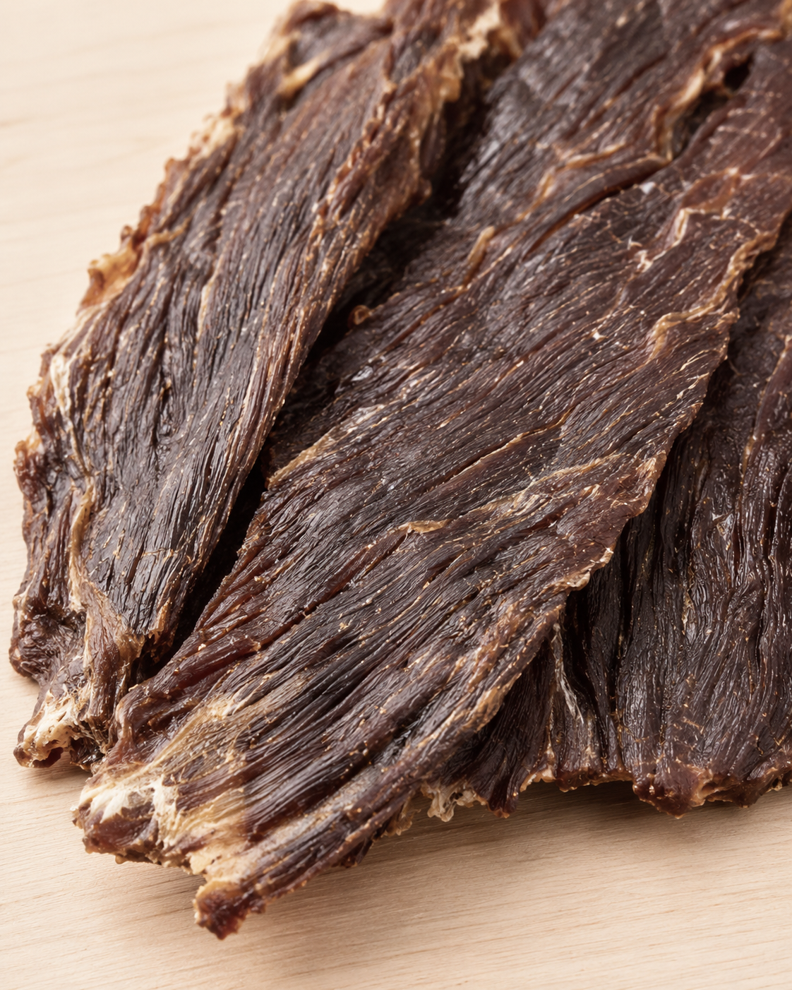Close-up of several strips beef jerky with visible texture, arranged on a light wood surface.